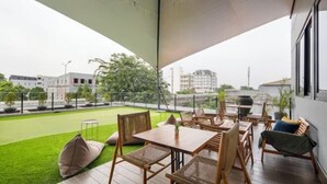A modern outdoor patio with a wooden table and chairs, a small lawn, and a view of the cityscape. - Cove Amartha (South Jakarta)