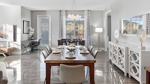 A modern dining area with a long table, chairs, and a chandelier. There is a kitchen area with a stove and cabinets visible in the background. - Modern 4BR Resort Townhome near Universal (Orlando)