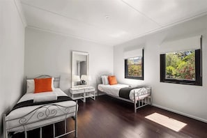 A bedroom with two single beds, a bedside table with lamps, a mirror, and a window with a view of greenery.