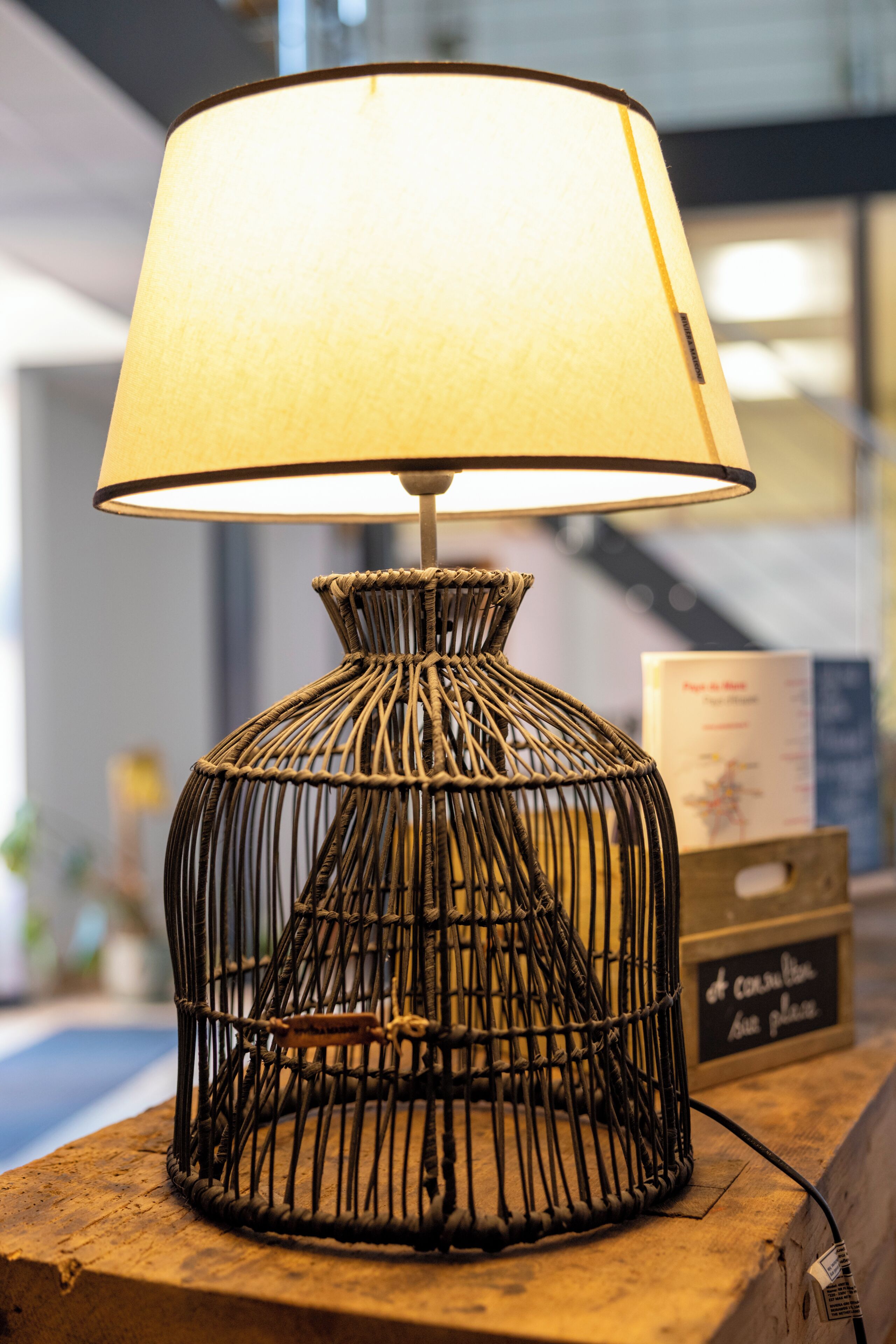 En lampe med flettet fod og hvid skærm på en træoverflade.