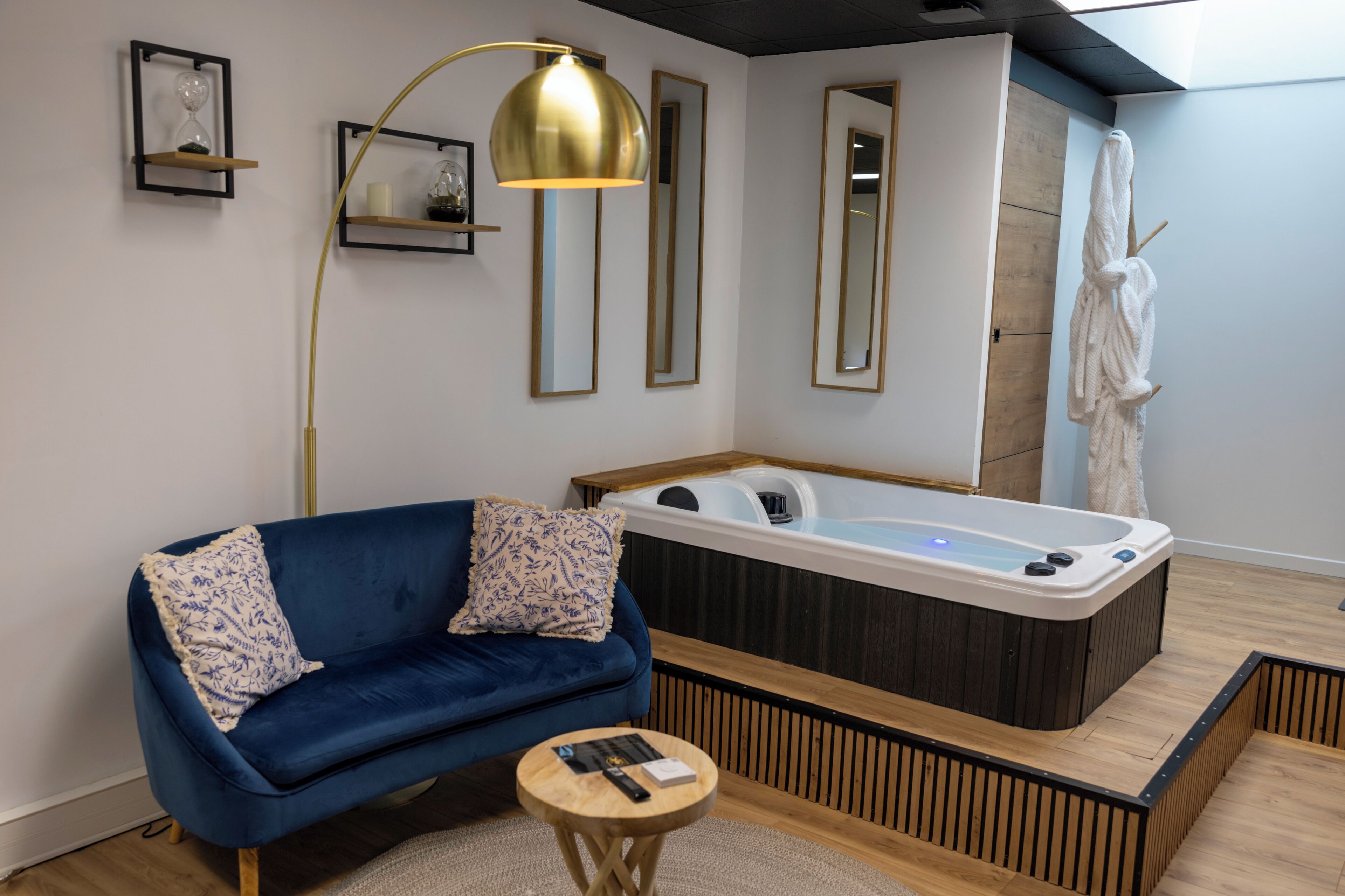 A modern room with a blue sofa, a round wooden side table, a large gold floor lamp, a white bathtub, and a white robe hanging on a hook.
