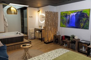 A modern hotel room with a large bathtub, a wooden floor, a round mirror, a wooden partition, a television, and a small table with a remote control. - Château de Posset - Les lofts (Teloché)
