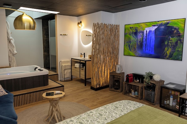 Una habitación de hotel moderna con una bañera grande, piso de madera, un espejo redondo, un panel de madera, un televisor y una pequeña mesa con un control remoto.