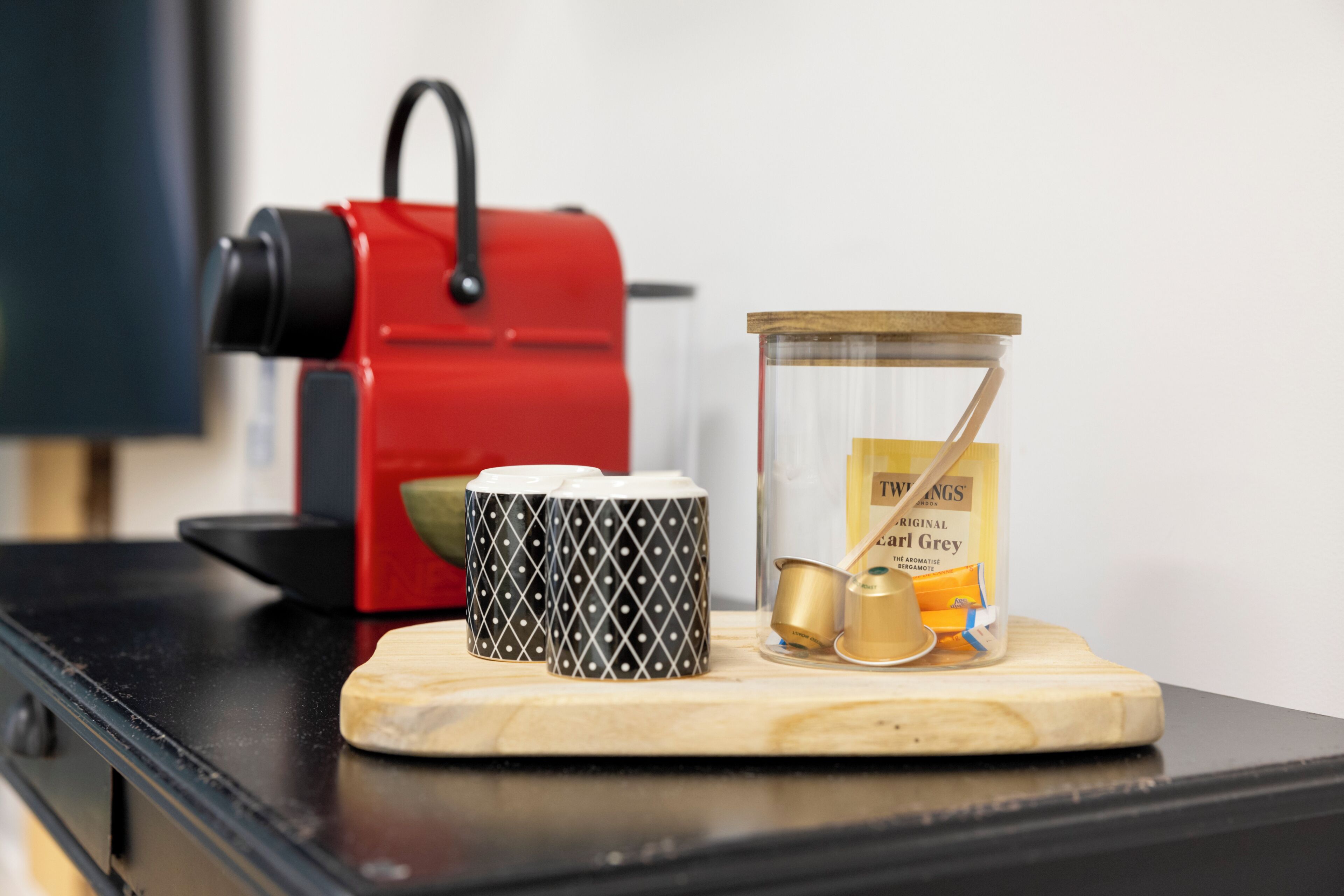 Une machine à café, une tasse et un récipient en verre contenant des sachets de thé, le tout posé sur une planche en bois.