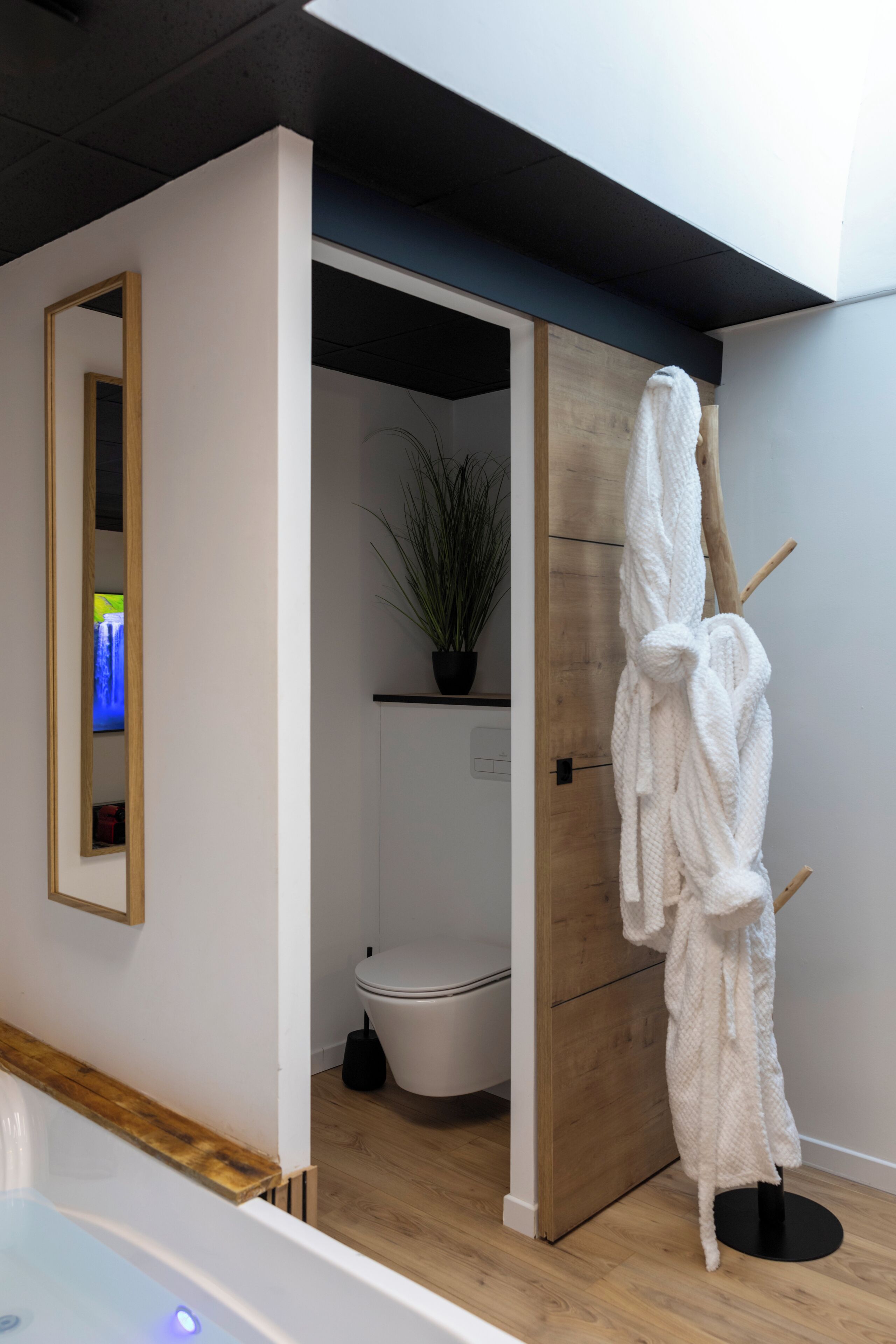 A modern bathroom with a wooden door, a white toilet, a potted plant, and a towel rack with a towel.