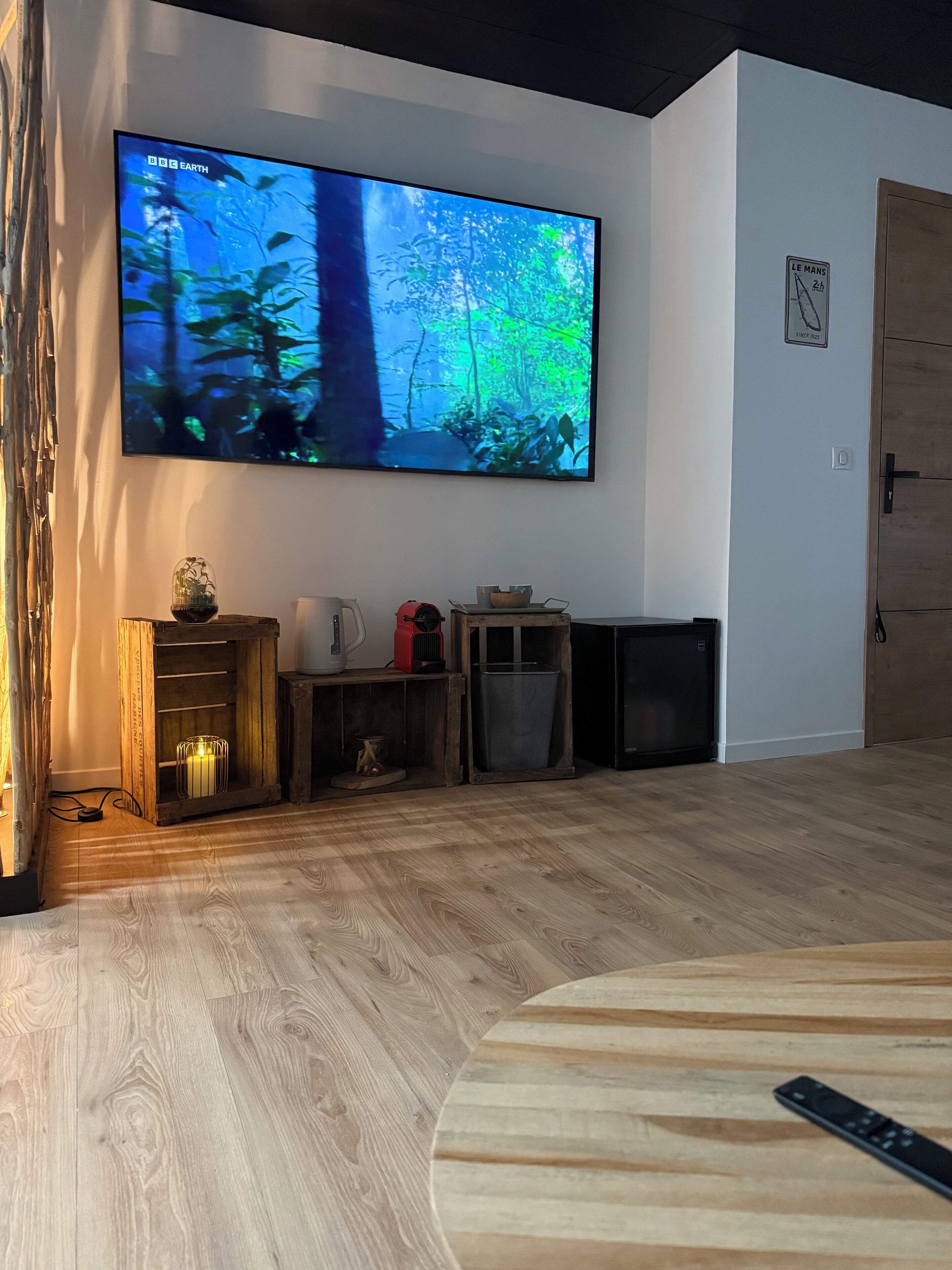 A living room with a large flat-screen TV mounted on the wall displaying a nature scene. There is a wooden cabinet with a small table and a lamp, and a door with a sign on it.