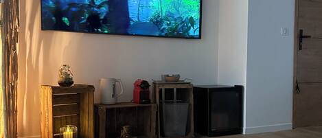 A living room with a large flat-screen TV mounted on the wall displaying a nature scene. There is a wooden cabinet with a small table and a lamp, and a door with a sign on it.