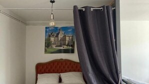 Un dormitorio con cabecera capitonada, cama con ropa de cama blanca, lámpara colgante y cortina oscura entreabierta.