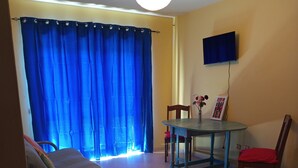 A room with a bed, a dining table with chairs, a TV mounted on the wall, and a window with blue curtains. - Gianni Apartment, Santa Maria, Sal, Cape Verde (Santa Maria)