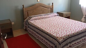 A bedroom with a wooden bed, a red rug, a nightstand, a wall-mounted painting, and a window with curtains. - Gianni Apartment, Santa Maria, Sal, Cape Verde (Santa Maria)