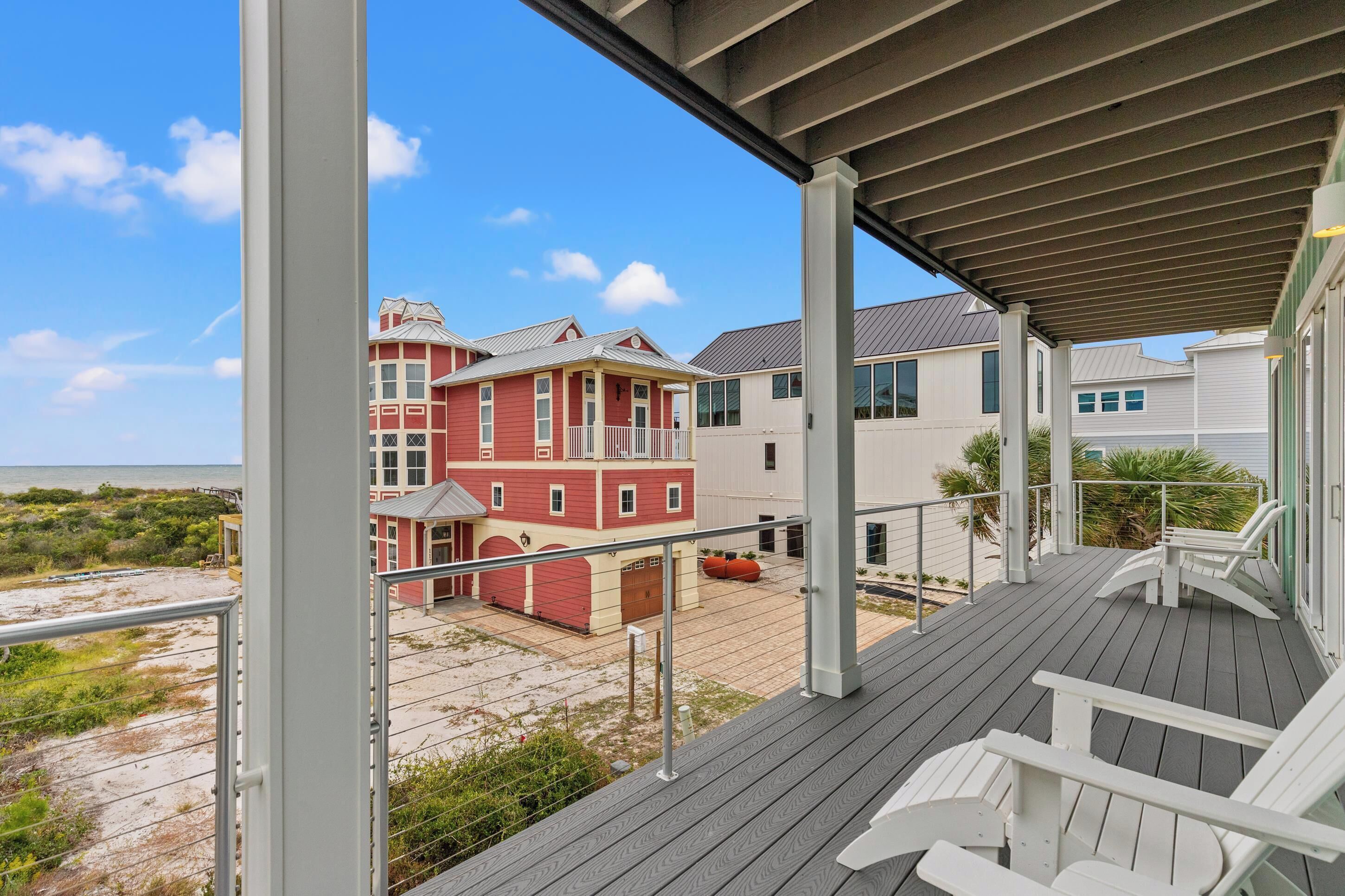 A covered deck with white chairs, a view of a red building, and a sandy beach area.