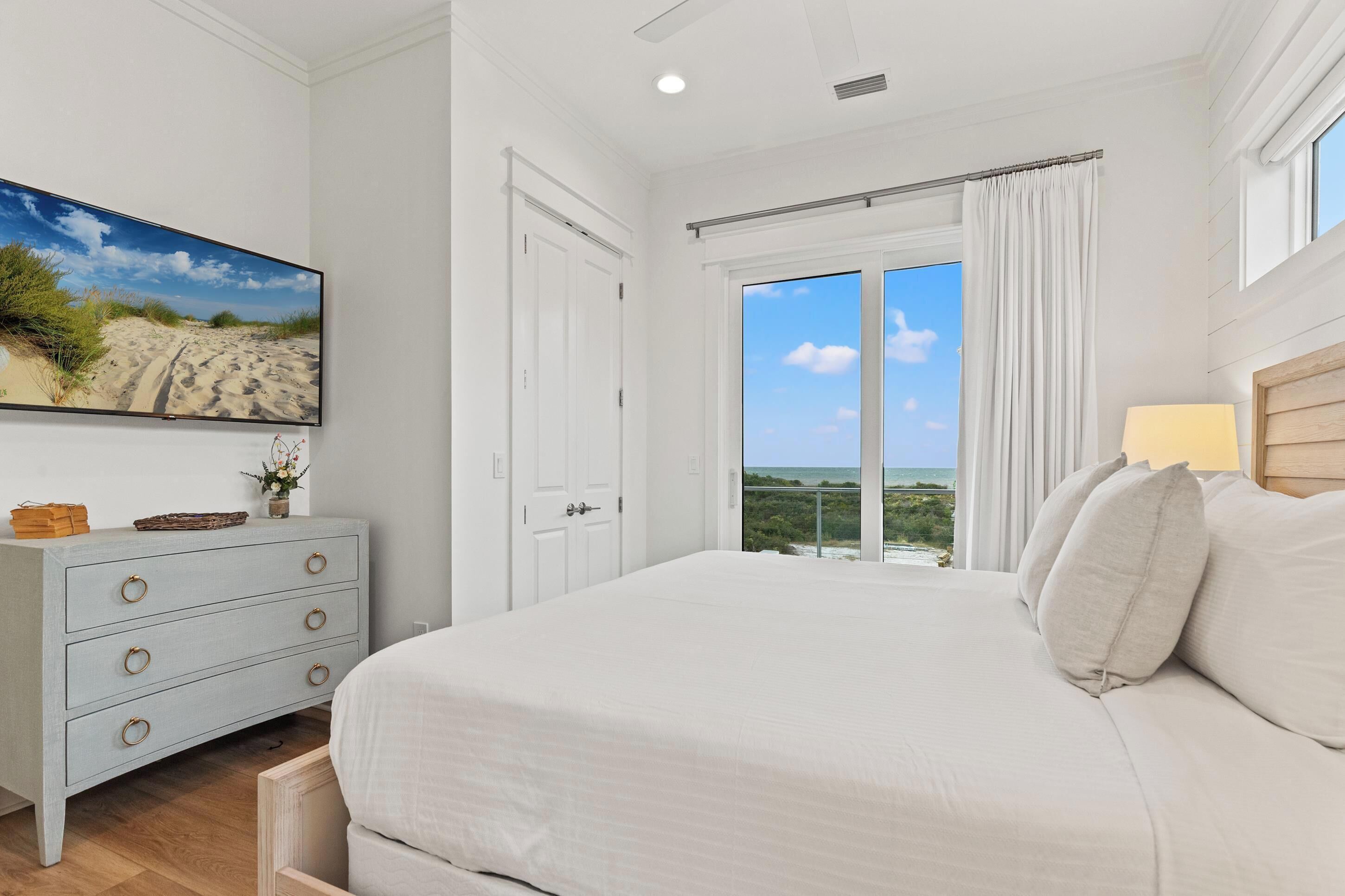 A bedroom with a large bed, a dresser, a painting of a beach, and a view of the ocean through a sliding door.