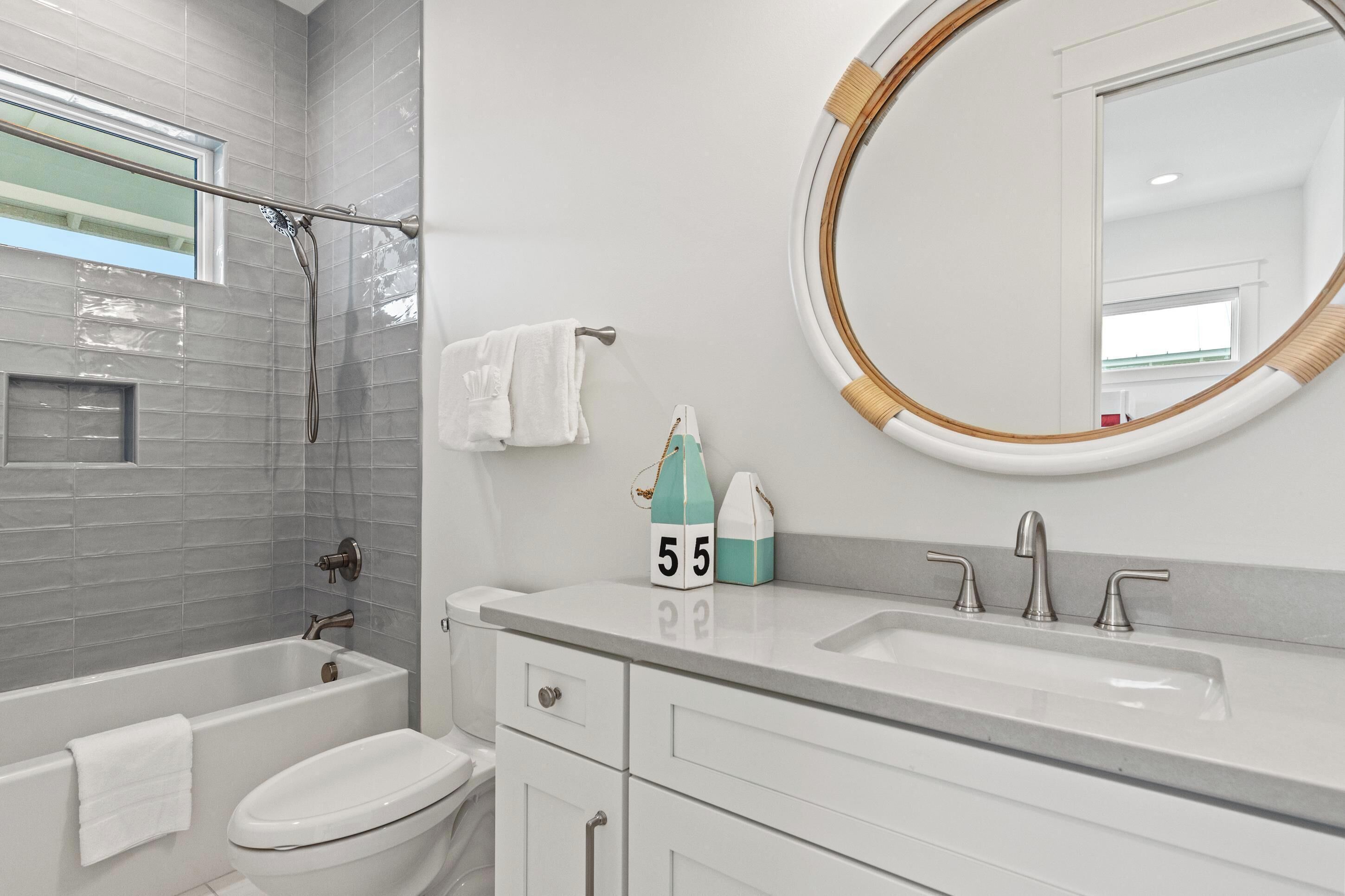 Une salle de bain moderne avec une baignoire, des toilettes et un lavabo. Il y a une fenêtre au-dessus de la baignoire et un miroir rond au-dessus du lavabo.