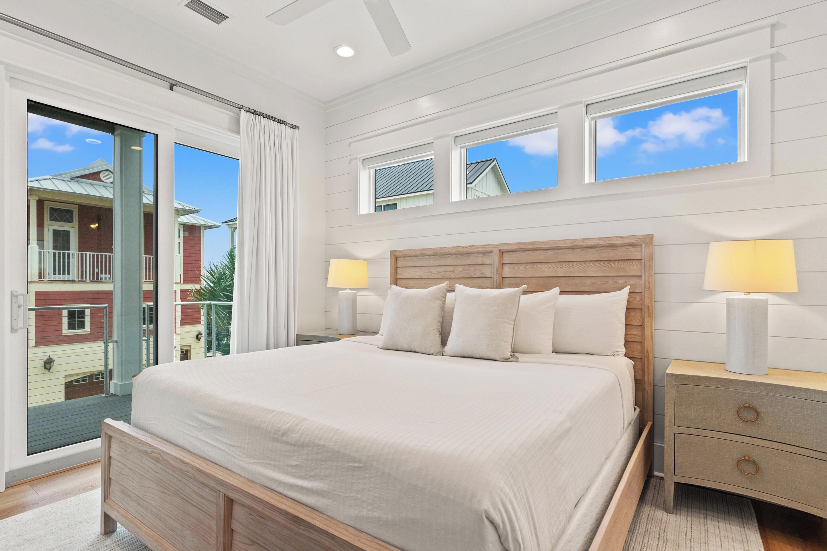 Quarto com uma cama grande, duas luminárias de cabeceira, um painel de cabeceira de madeira e vista para um edifício vermelho através de uma porta de vidro deslizante.