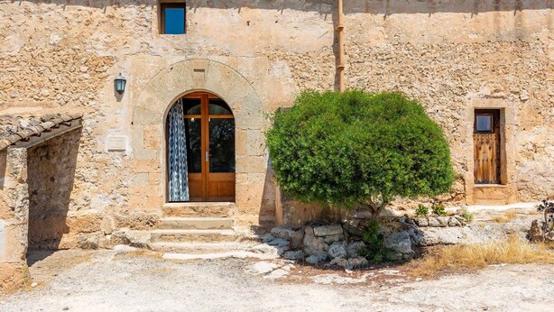 A rustic stone building with a wooden door, a small window, and a tree in front. - Can Gallot De Punxuat – Mountain Views, Private Pool, Wi-Fi & Air Conditioning (Algaida)