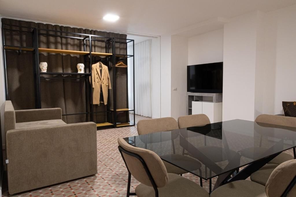Habitación de hotel moderna con una mesa de comedor de vidrio, sofás color beige, un armario metálico negro y un televisor de pantalla plana.