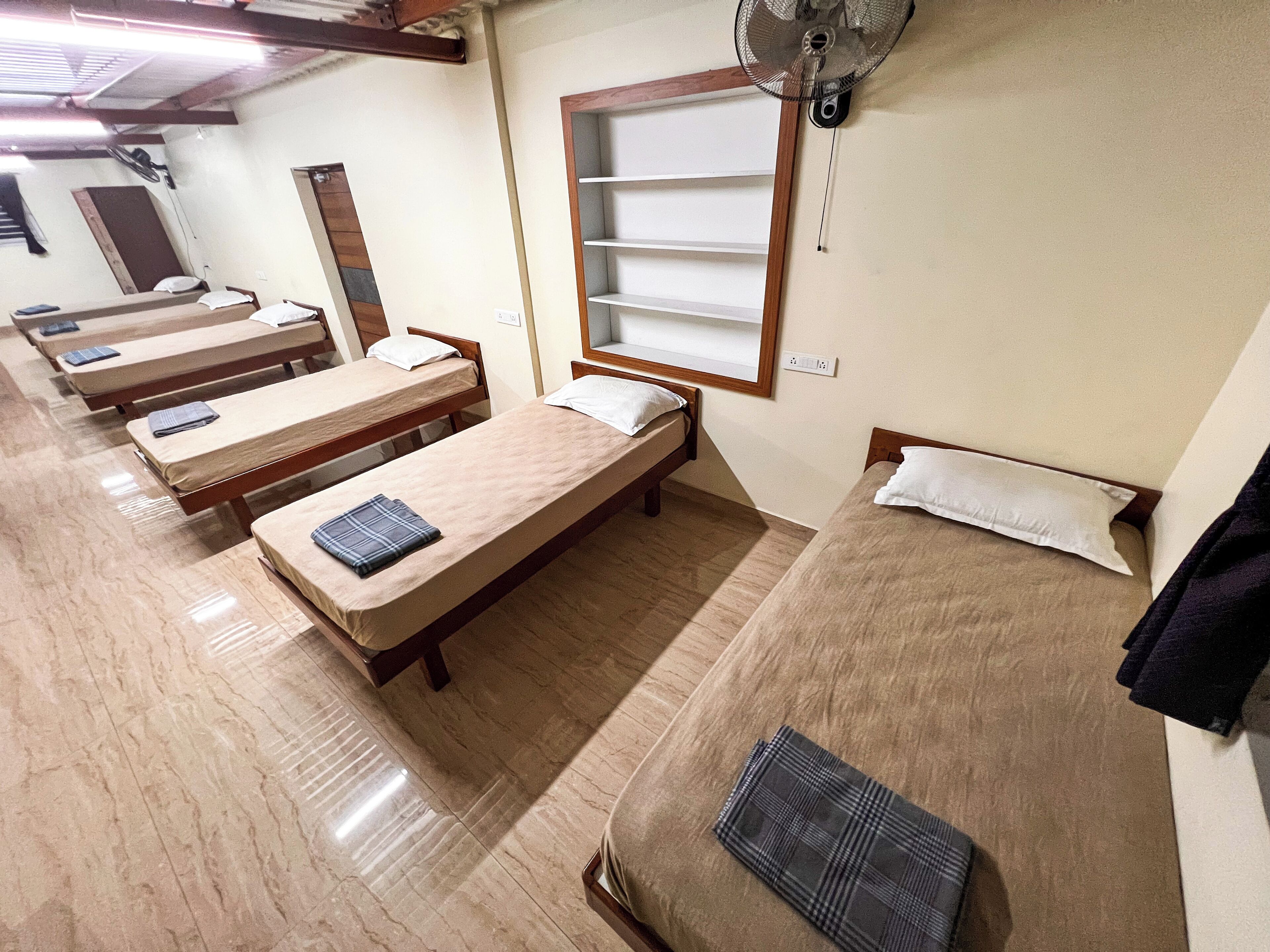 A room with four single beds, each with a pillow and a blanket. There is a window with a shelf and a wall-mounted fan.