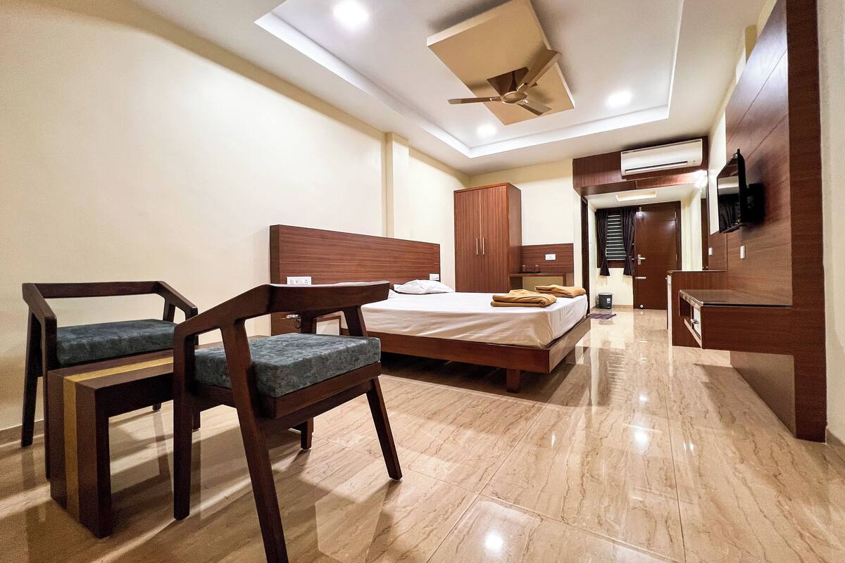 A modern hotel room with a bed, two wooden chairs, a desk, a ceiling fan, and a TV mounted on the wall.
