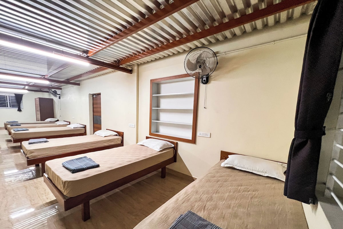 A dormitory room with four single beds, a standing fan, a window with shelves, and a door.