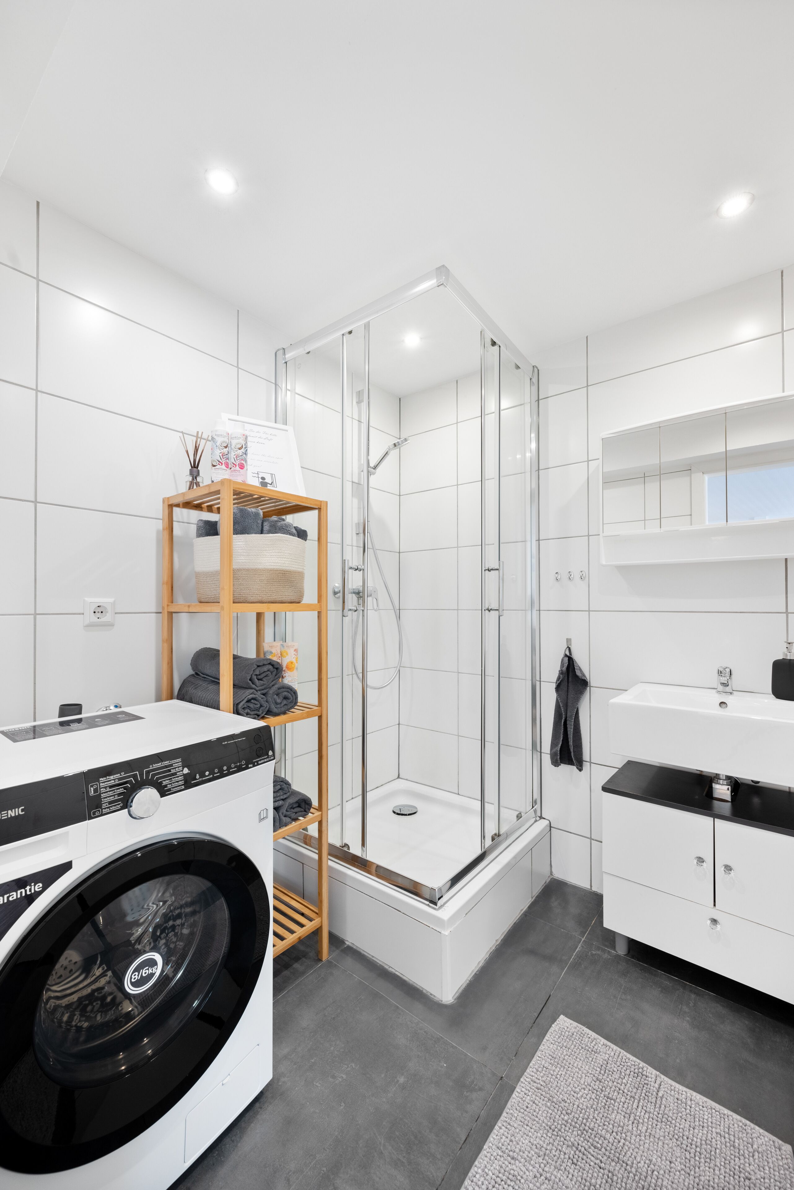 A modern bathroom with a glass-enclosed shower, a built-in washing machine, and a white vanity with a mirror.