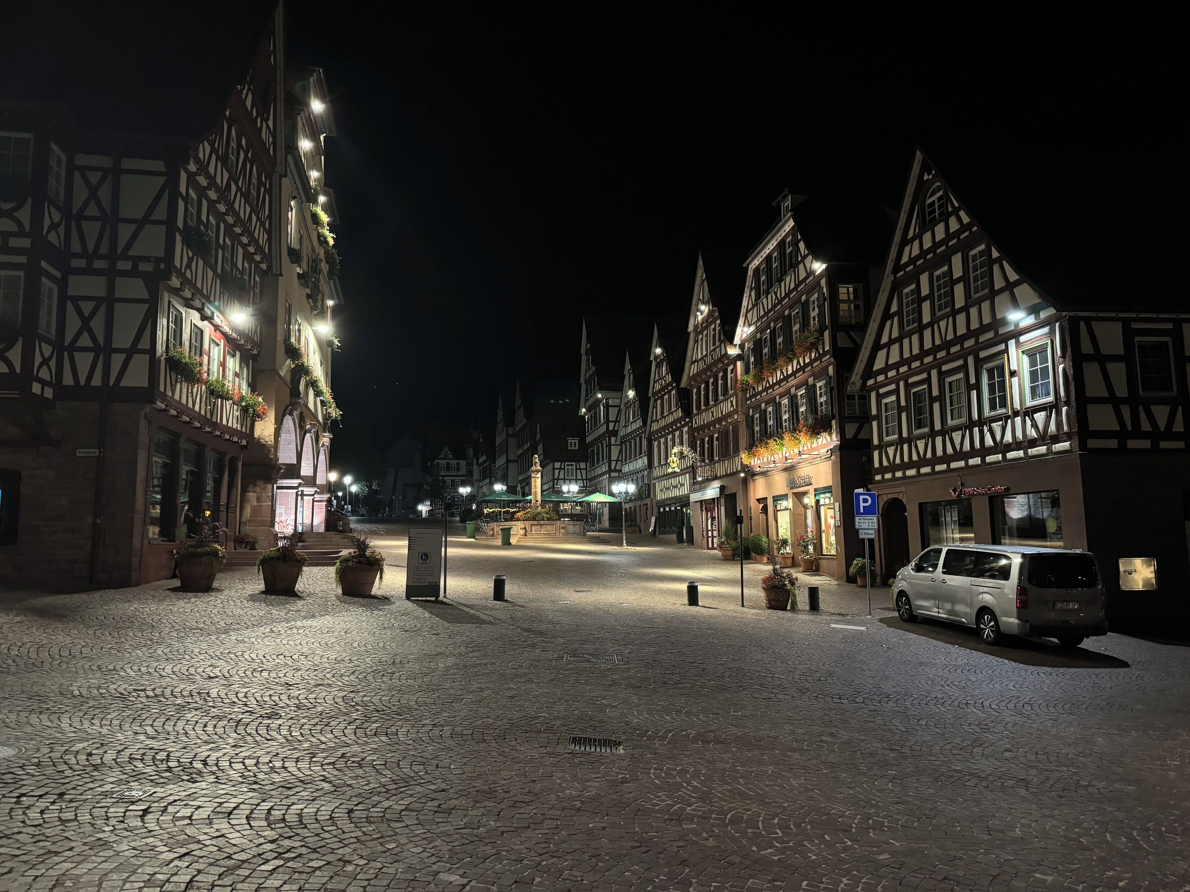 A cobblestone street lined with traditional half-timbered buildings, a central square with a fountain, and a parked van.