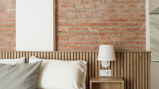 A bed with white linens and a grey pillow, a wooden bedside table with a white lamp, and a brick wall. - Guest Ready - Fira Barcelona Modern Studio (L'Hospitalet de Llobregat)