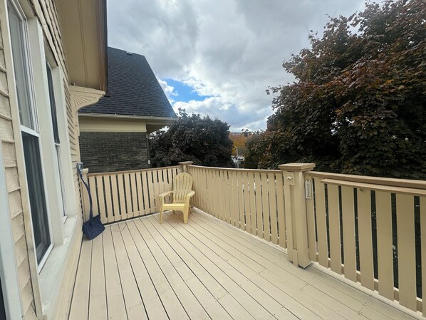 Une terrasse en bois avec une rambarde beige, une seule chaise et une vue sur un bâtiment en briques et des arbres.