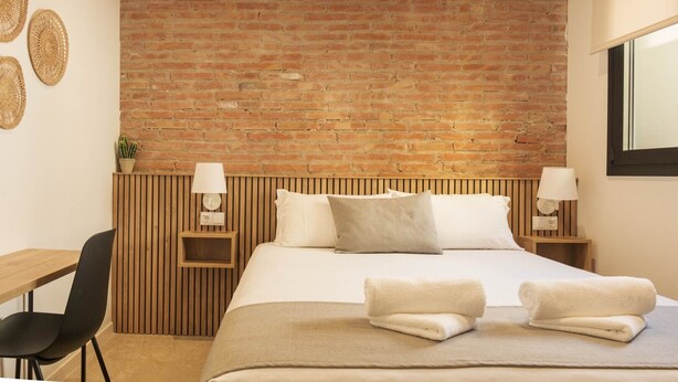 Hotelkamer met een bakstenen muur, een houten hoofdbord, een bed met witte lakens, twee nachtkastjes met lampen, een bureau met stoel, en een potplant.