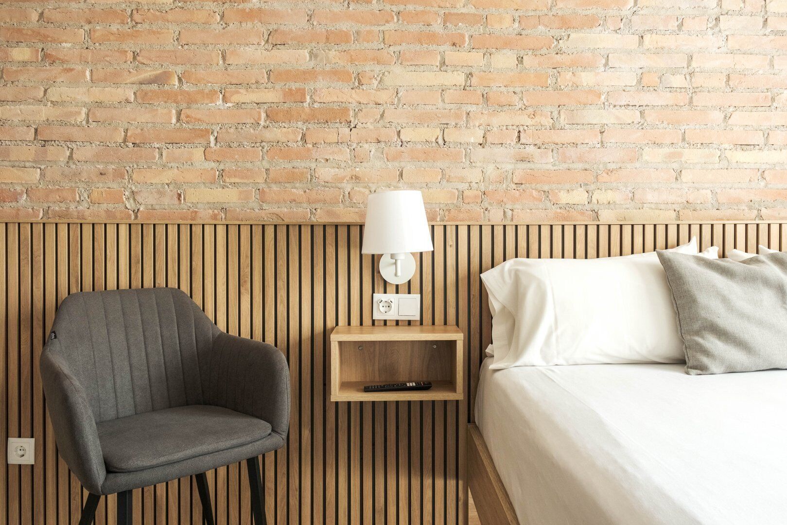 A hotel room with a brick wall, a wooden slatted headboard, a grey armchair, a bedside table with a lamp, and a bed with pillows.