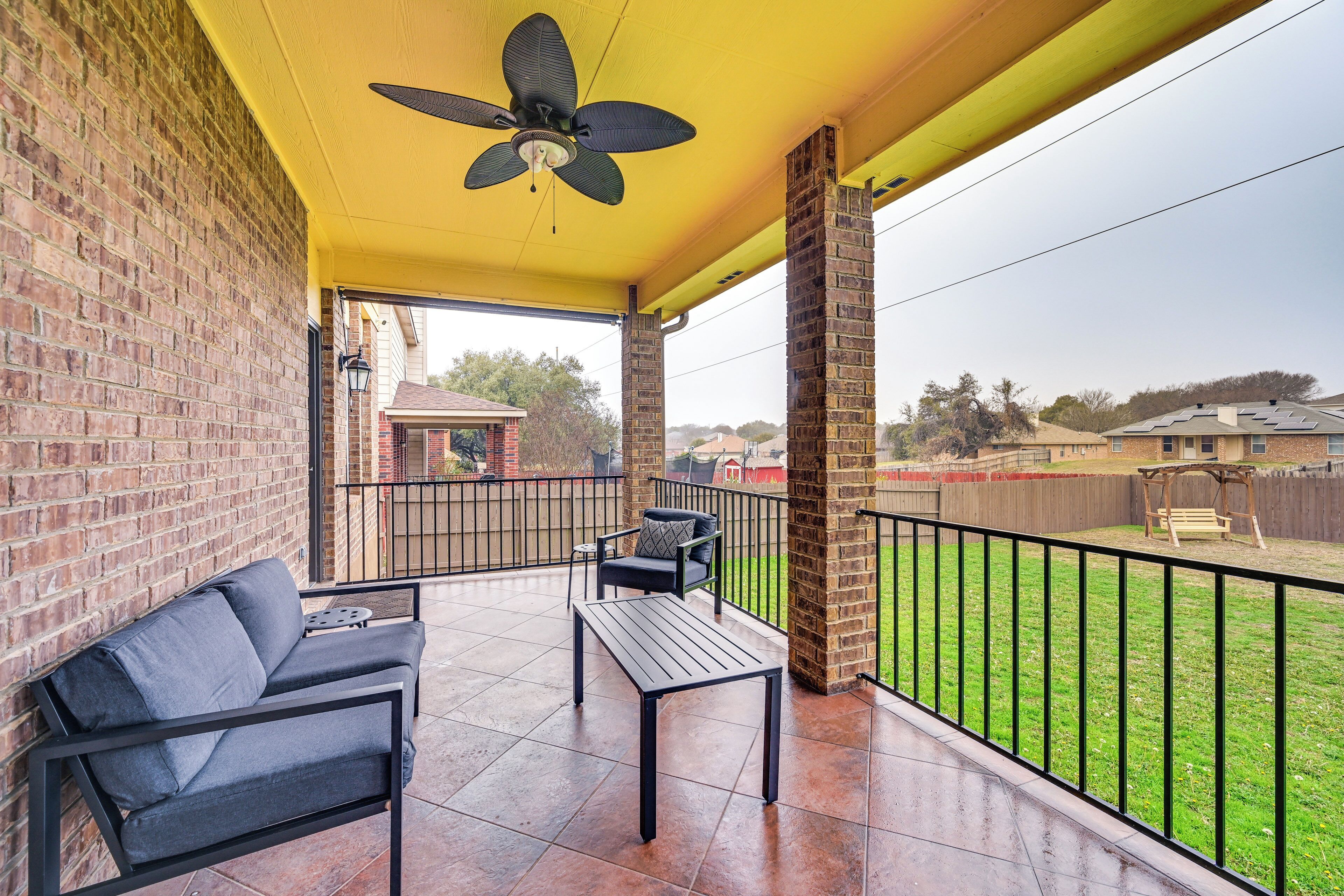 A covered patio with a brick wall, a ceiling fan, a black metal table and bench, and a view of a backyard with a fence and trees.