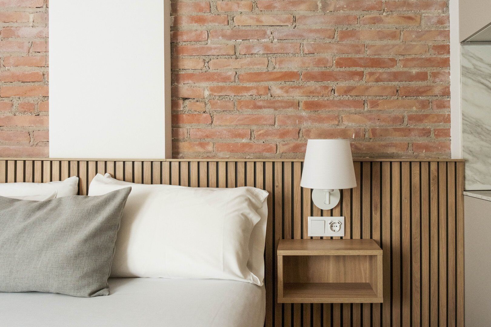 Una cama con sábanas blancas y un almohadón gris, una mesita de noche de madera con una lámpara blanca, y una pared de ladrillo.