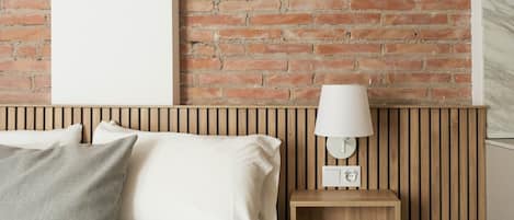 Una cama con sábanas blancas y un almohadón gris, una mesita de noche de madera con una lámpara blanca, y una pared de ladrillo.