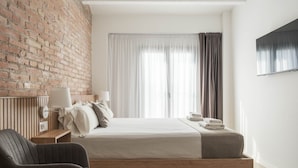 Een moderne hotelkamer met een bakstenen muur, een bed met witte lakens, een nachtkastje met een lamp en uitzicht op de stad door dunne gordijnen.