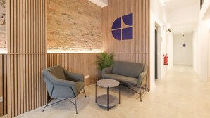 A modern lobby with a brick wall, wooden slats, two grey armchairs, a small round table, and a large logo on the wall. - Guest Ready - Fira Barcelona Modern Studio (L'Hospitalet de Llobregat)