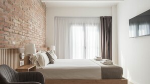 A modern hotel room with a brick wall, a bed with white linens, a bedside table with a lamp, and a view of the city through sheer curtains. - Guest Ready - Fira Barcelona Modern Studio (L'Hospitalet de Llobregat)