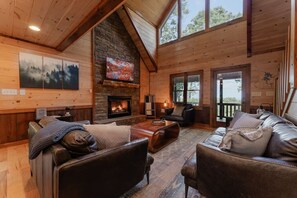 A cozy cabin interior with a fireplace, leather sofas, and a large window. - Mountain Memories:HotTub, GameRoom-KingSuites (Blue Ridge)