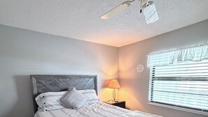 Quarto com cama, mesa de cabeceira, abajur, ventilador de teto e janela com persianas.