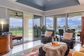 A spacious living area with a large sliding glass door leading to a balcony, a television, a fireplace, and a dining area with chairs and a table. - Tropical Elegance 2BR Condo; Beach/Golf/Shopping (Waikoloa Village)