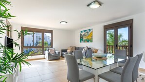 A modern living room with a glass dining table, grey chairs, a sofa, and a painting on the wall.