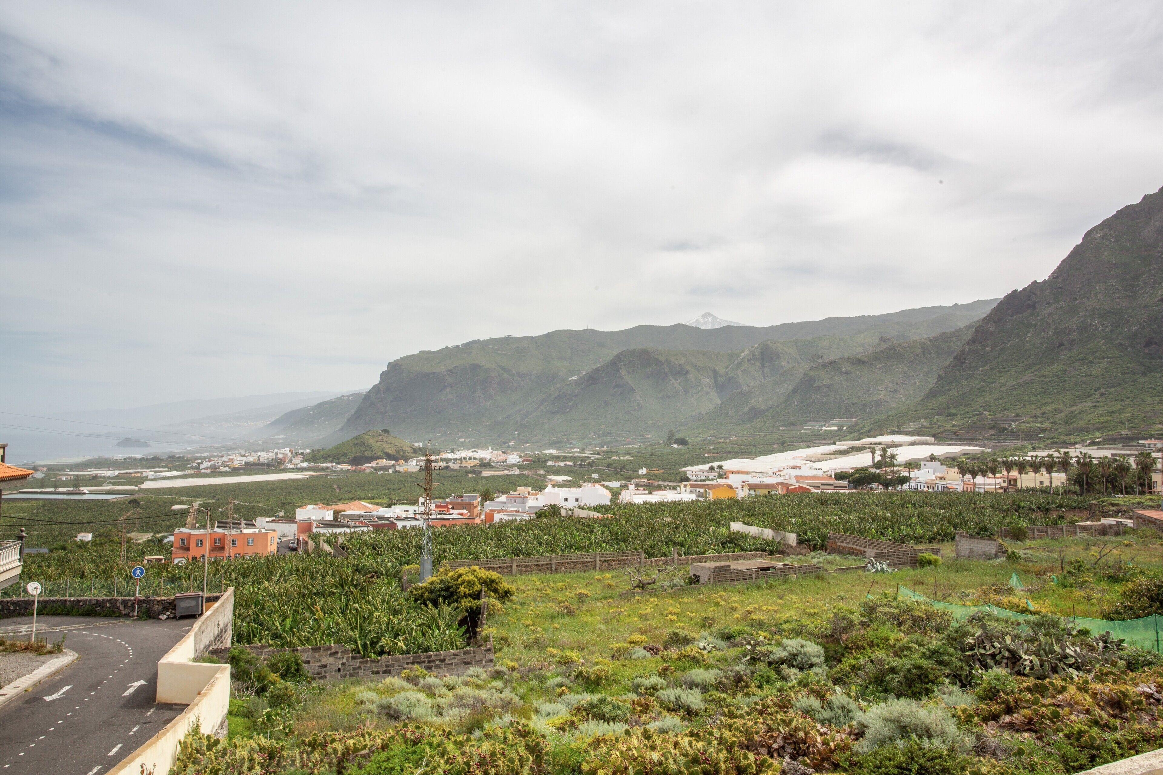A coastal town with a mountain backdrop, a road with a blue sign, and buildings with red and white facades.