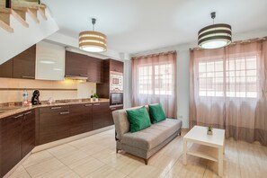 Een moderne keuken met donkere kasten, een grijze bank met groene kussens en een witte tafel met een plant.