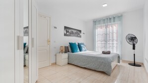 Une chambre à coucher comprenant un lit, une table de chevet, une commode, des œuvres d’art murales, une fenêtre avec des rideaux et un ventilateur sur pied.