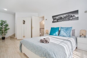 Een slaapkamer met een bed, een kamerplant, een deur, een aan de muur hangend schilderij en een nachtkastje.