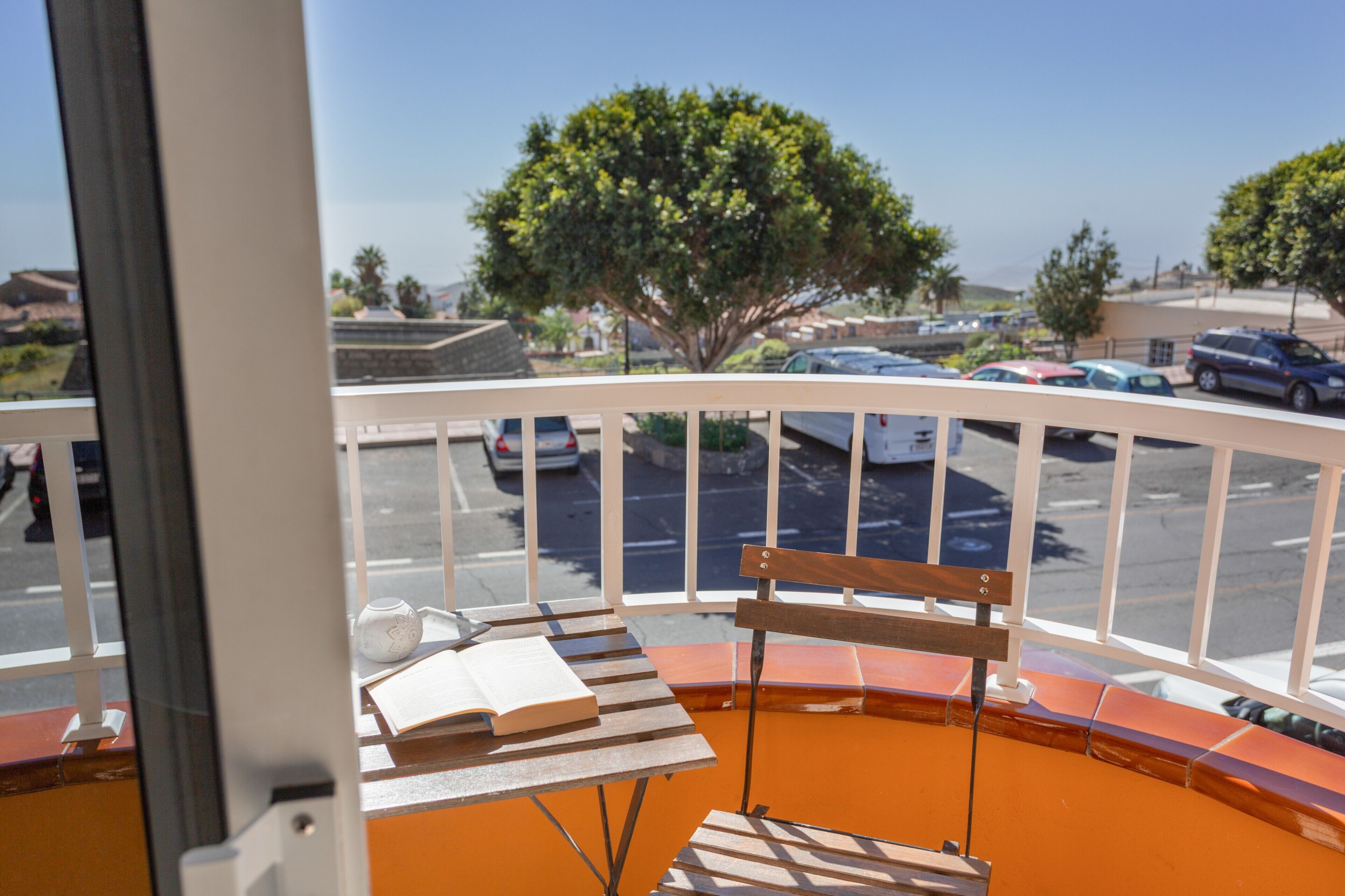 Un balcon avec un banc en bois, une table avec un livre, et une vue sur un parking et des arbres.