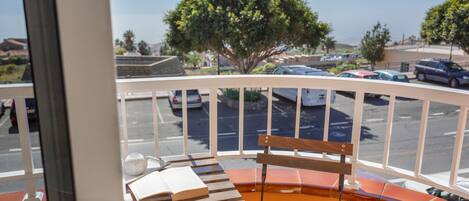 Un balcon avec un banc en bois, une table avec un livre, et une vue sur un parking et des arbres.
