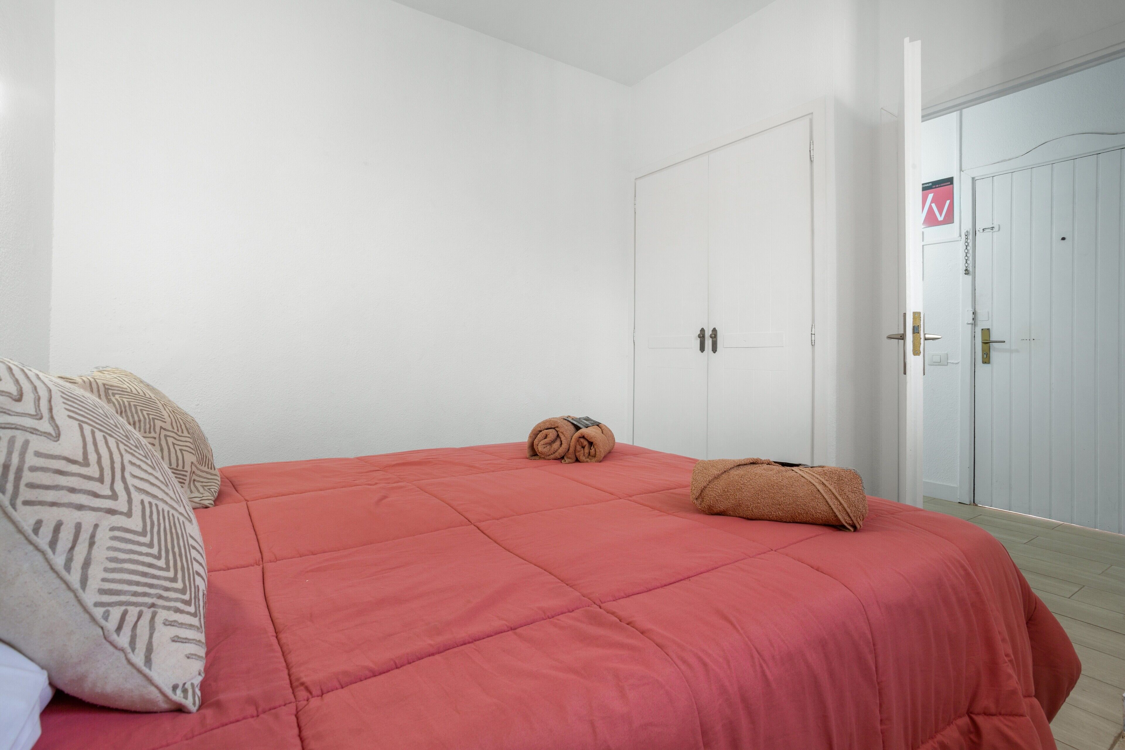 A bedroom with a red bedspread, two pillows, a rolled towel, and a white door with a sign.