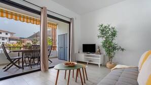 Ein modernes Wohnzimmer mit einer Couch, einem Fernseher, einem Esstisch mit Stühlen, einem Balkon mit Aussicht und einer Topfpflanze.