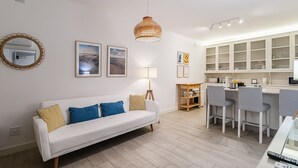 Un soggiorno moderno con un divano bianco, una zona pranzo con sgabelli da bar, una cucina con mobili bianchi e un lampadario in rattan.