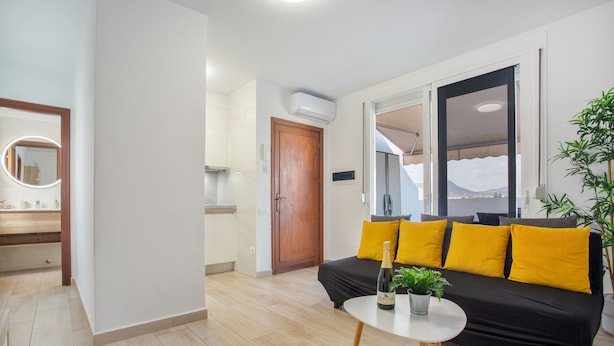 A modern living room with a black sofa, yellow cushions, a small round table with a bottle and plant, and a wooden door.