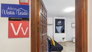 A hotel interior with a wooden door, a blue sign with white text, a red sign with a white letter, a black sofa with cushions, a small table with a plant, and a wall-mounted picture.